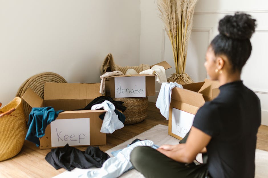 A woman with dark curly hair tied up in a bun is sitting on a light wooden floor in a bright, minimally decorated room. She is facing to the right and is dressed in a black short-sleeved top. Around h