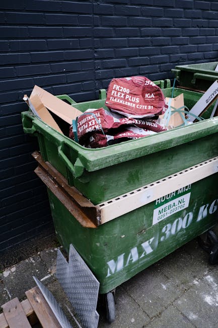 A large green wheeled waste bin positioned outdoors on a paved surface next to a dark brick wall. The lid of the bin is open, revealing several red and purple bags filled with construction or garden w
