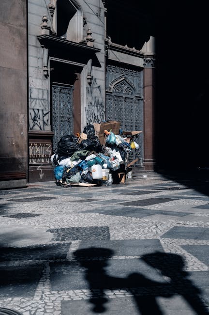A large green wheeled waste bin positioned outdoors on a paved surface next to a dark brick wall. The lid of the bin is open, revealing several red and purple bags filled with construction or garden w